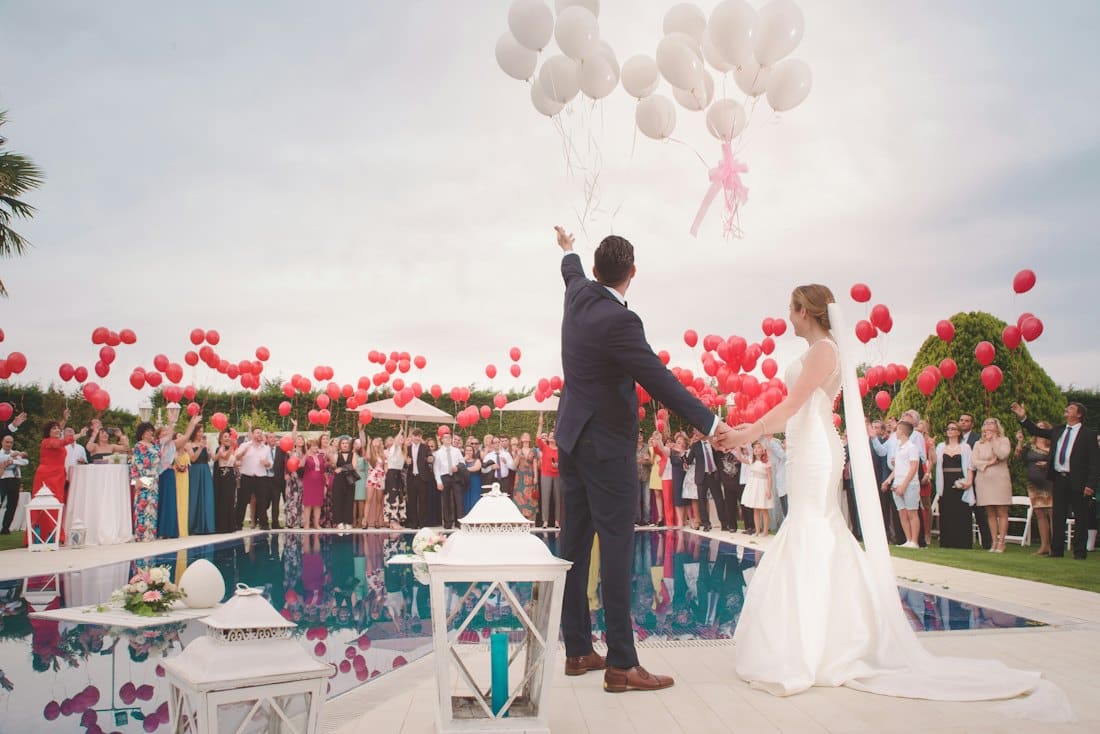 Newlyweds raising their hands beside a poolside venue decorated with red and white balloons