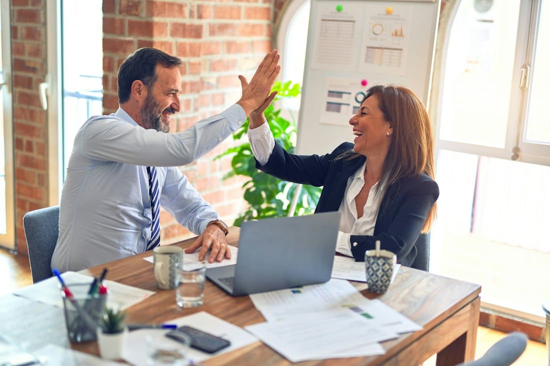 Two consultants high-fiving over a desk of laptops and printed reports