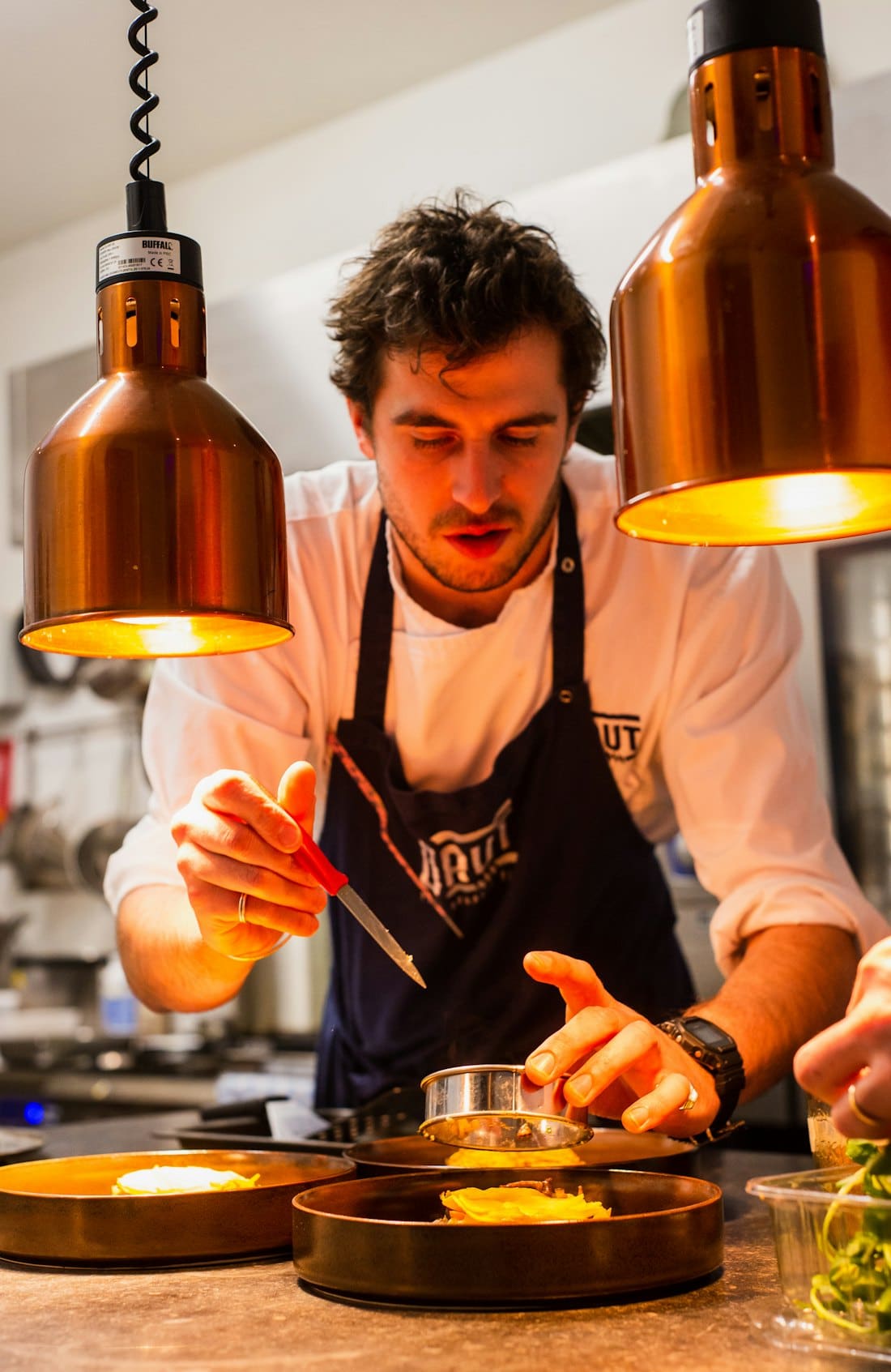 Head chef in an apron plating dishes under copper heat lamps